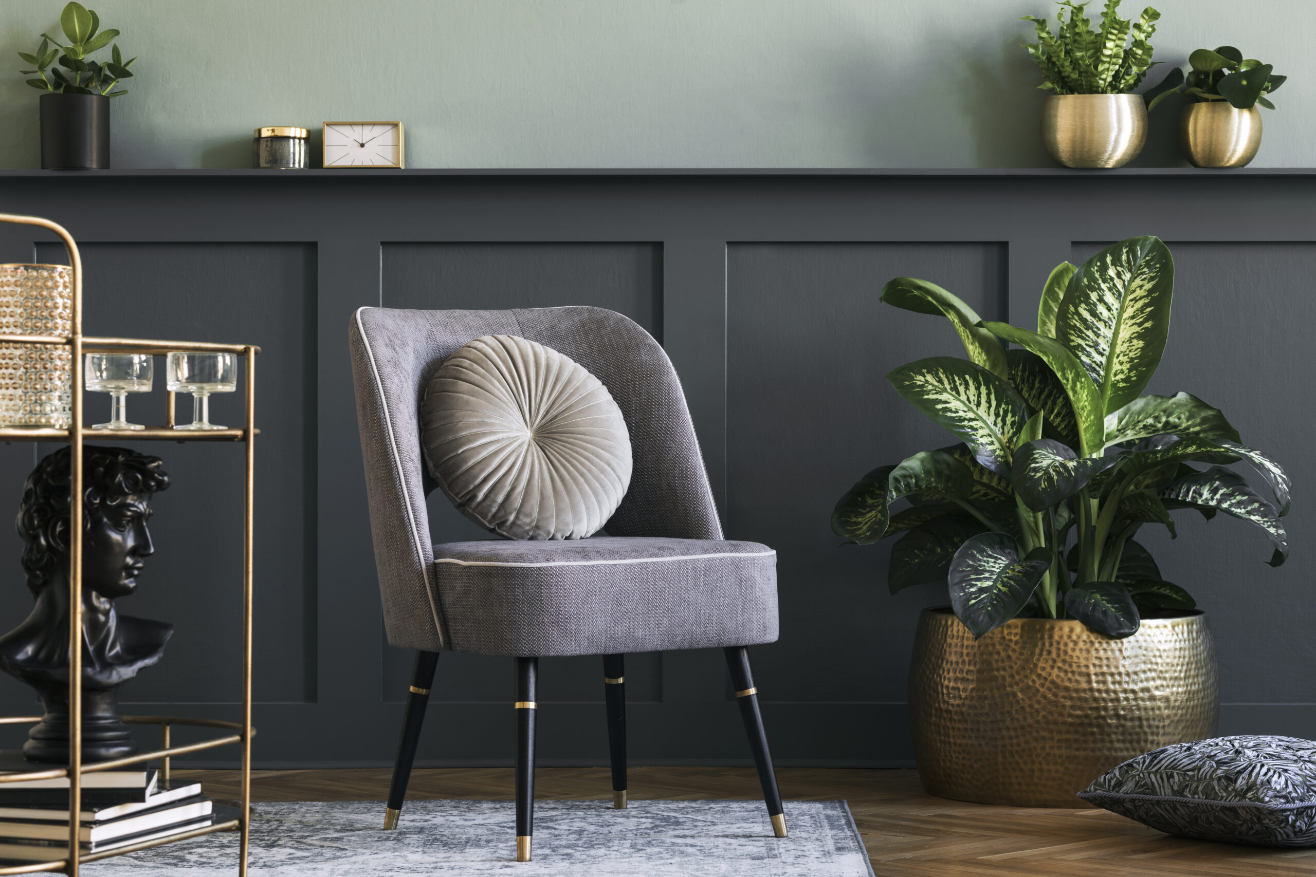 Stylish and modern composition of living room with design gray armchair, gold liquor cabinet, plants and elegant personal accessories. Gray wall panelling with shelf. Modern home decor. Template.